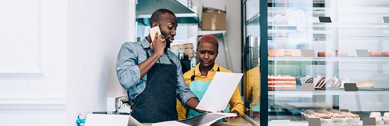 Male and female entrepreneur standing in front of shelving while wearing aprons. Reviewing their executive summary to check for any mistakes.