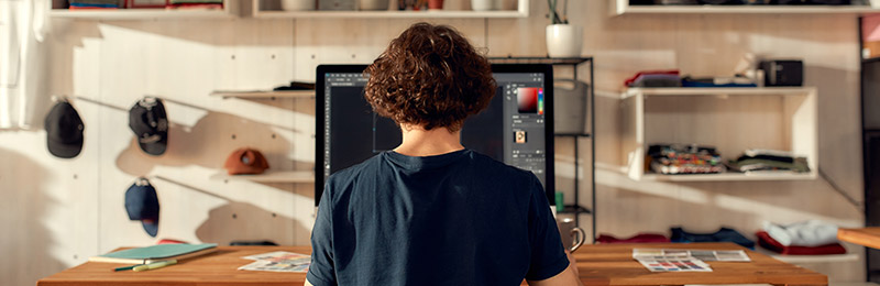 Male fashion entrepreneur sitting at desk in front of a computer with various pieces of apparel surrounding him. Working on formatting the styling and structure of his business plan.