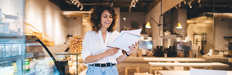 Female entrepreneur standing in the front of her bakery reviewing her business planning documents.