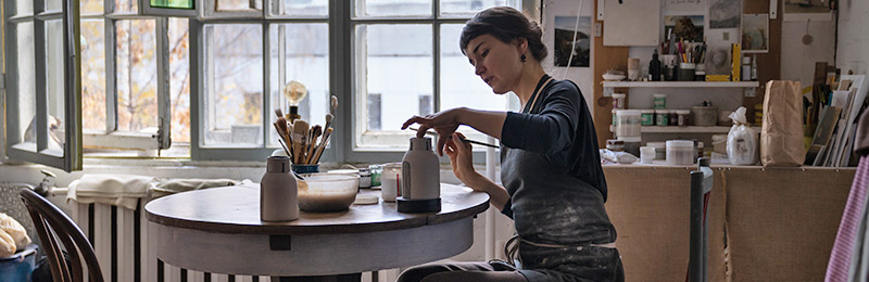 Female entrepreneur working on a piece of clay pottery within her shop. Considering if she needs to write a business plan for her art business.